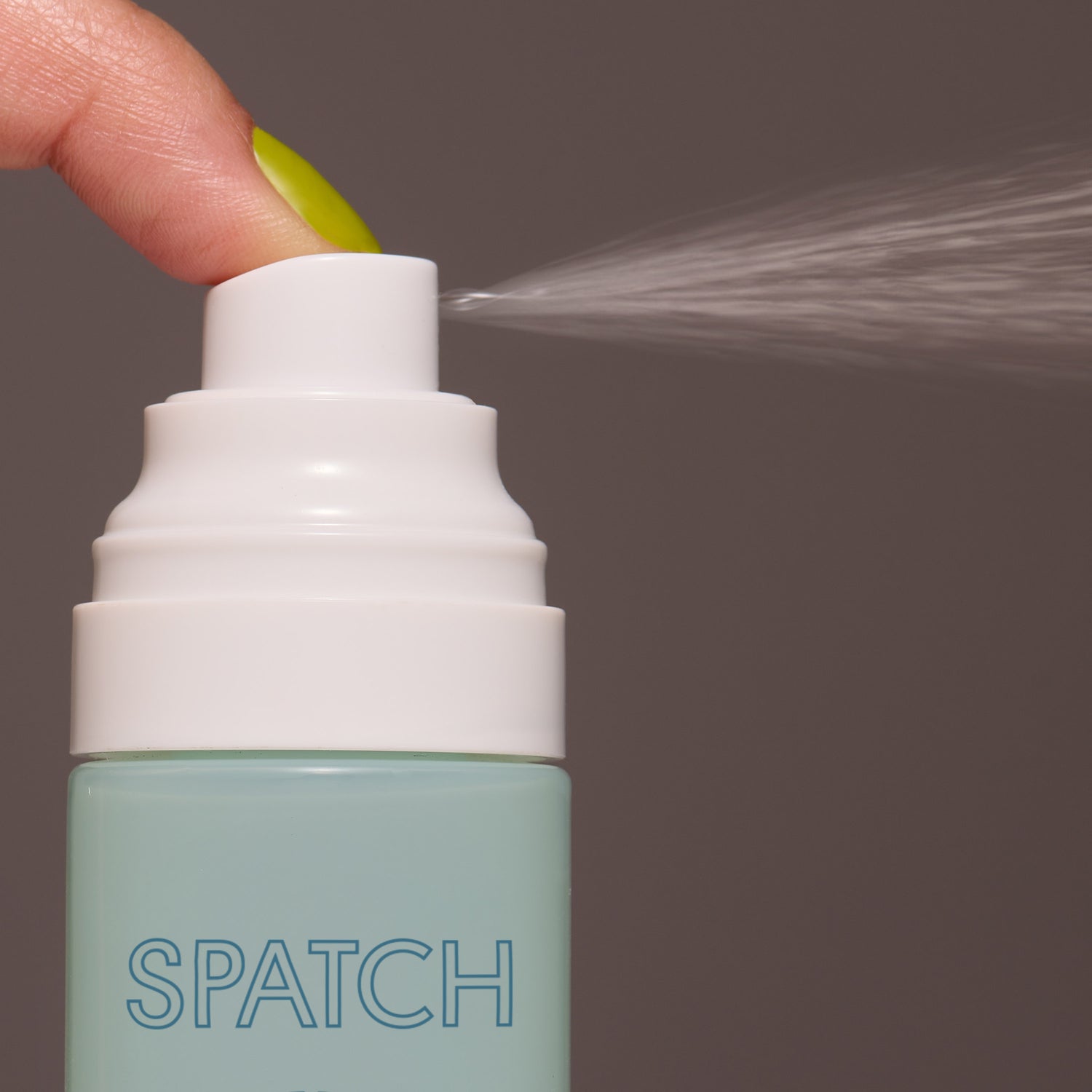 A finger with a bright yellowish green nail presses down on a bottle of makeup setting spray, causing the liquid to spray out of its mint/aqua green bottle. 