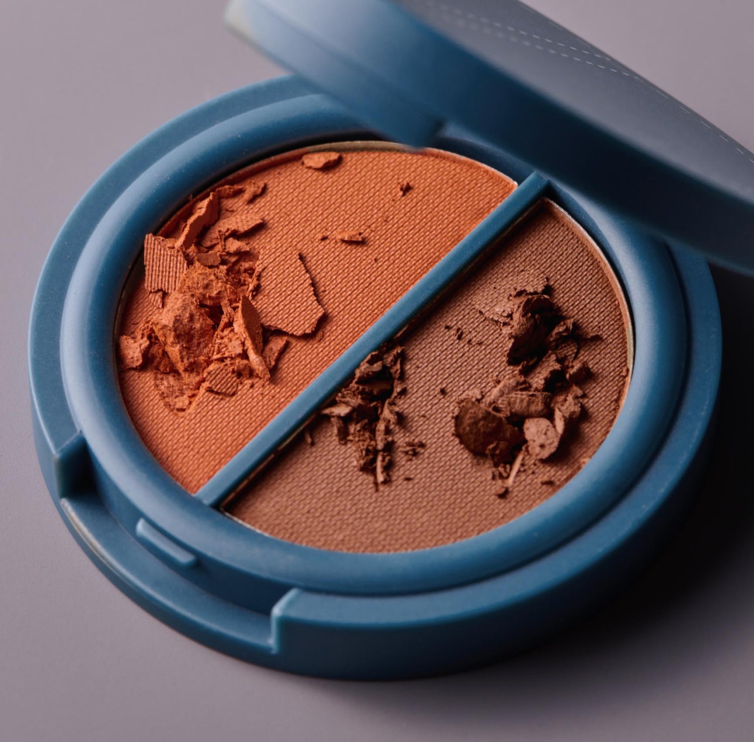 A round, teal-colored compact of brown pressed cosmetic powder in two hues.